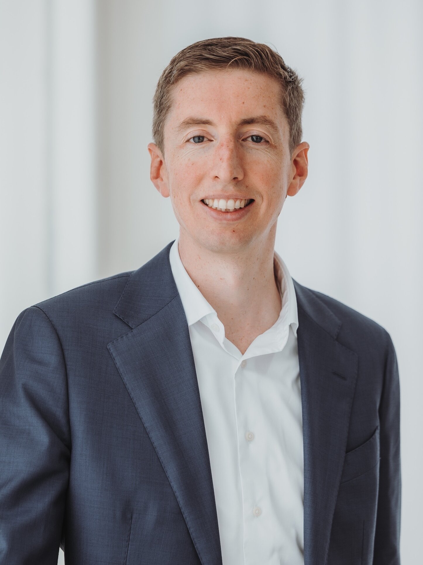 Portrait of a smiling man in a navy blazer and white shirt, standing indoors against a light curtain backdrop.